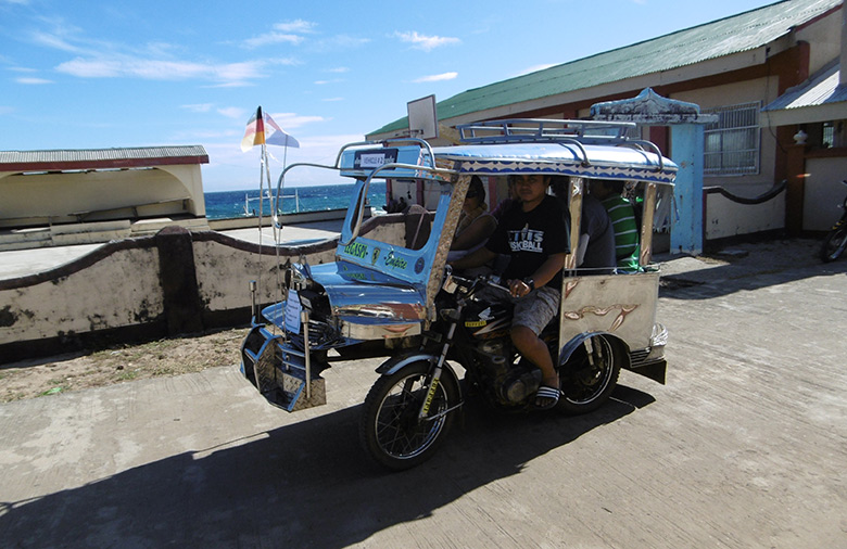 Das Trcycle, eine Art überdachtes Moped, mit mehreren Passagieren ist zu sehen; im Hintergrund das Meer.